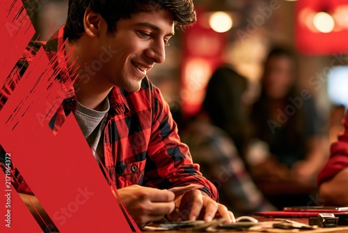 Young man wearing red plaid shirt playing card game with friends at indoor social gathering venue