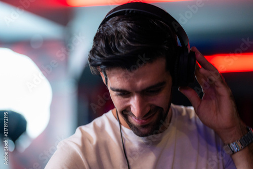 Foto A striking studio-style portrait of a smiling bearded DJ wearing black headphones holds one headphone cup with his hand and grins against a bright glowing club window