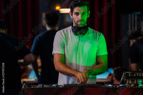 Obraz na plátně A wide view of a male DJ standing behind turntables and a mixer in a busy nightclub stands behind the turntables with the pool tables lit behind him in a warm candid medium shot