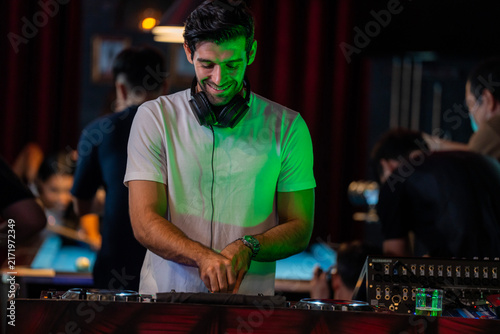 Obraz na plátne A wide view of a male DJ standing behind turntables and a mixer in a busy nightclub keeps both hands on the deck as guests move in the background in a warm candid medium shot