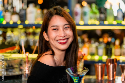 Fotografie A beautiful young Asian woman in a black off-shoulder top holding a bright blue cocktail smiles wide with the blue drink held at chest height in a flattering beauty close-up