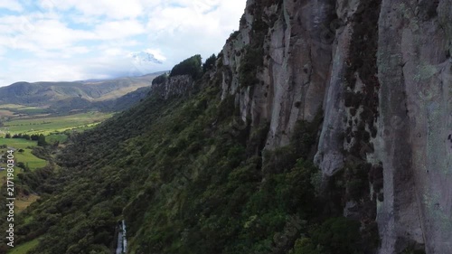 Aerial drone shot passing close to a tall rock cliff covered in moss and lichen, revealing a green farming valley and distant mountains in the Ecuadorian Andes.