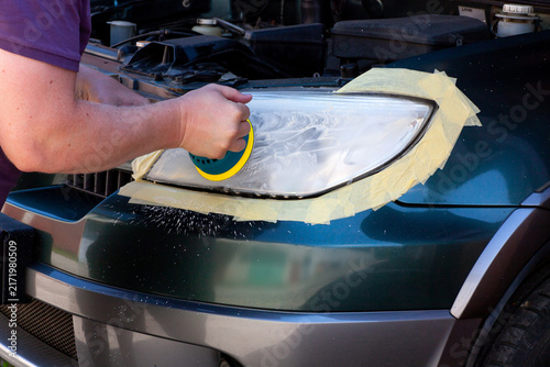 A person is seen polishing a car headlight in a garage. They use a tool and tape is applied around the headlight area for protection. The sun shines through the garage.