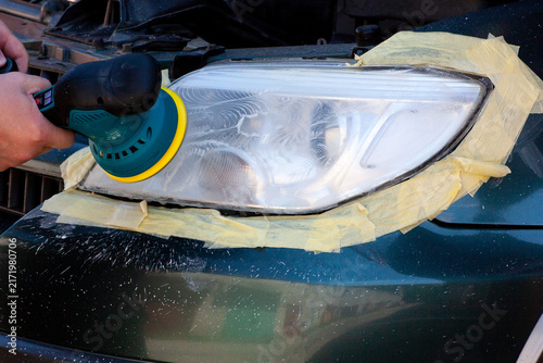 A person is using a polishing tool to clean a car headlight. The headlight is masked with tape. The setting is an auto workshop. Dust and debris are visible in the air.