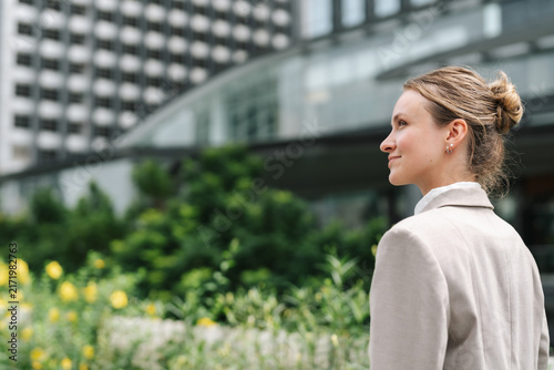 Profile portrait of young businesswoman with hair bun looking away outdoors, wearing beige blazer near office building and green garden, thoughtful professional lifestyle