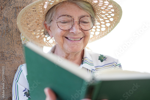 Canvastavla Smiling senior woman wearing a straw hat and round glasses enjoying a good book