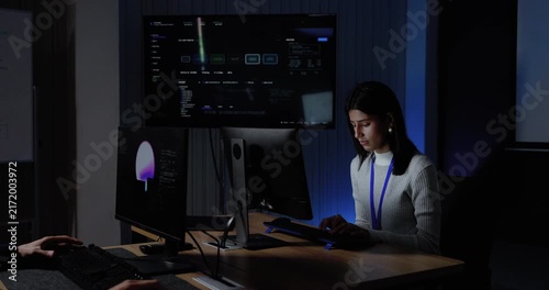 Adult female technology professional types at computer as adult male colleague approaches, leans beside workstation, and joins software dashboard review during collaboration inside modern office.
