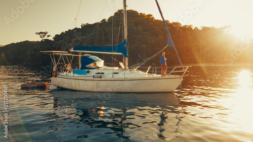 Man stands on the deck of the sailing boat and enjoys calm morning time in the secured lagoon on sunrise