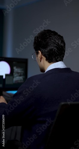 Young male professional works at computer workstation, reviewing software interface on monitor while making small head movements during system analysis, digital workflow, technology project planning.