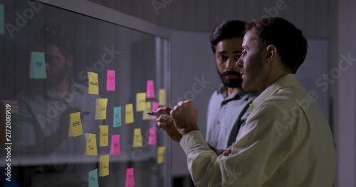 Two adult male technology professionals discuss sticky notes on glass board, pointing at project ideas and reviewing workflow during collaborative software strategy planning session in modern office.