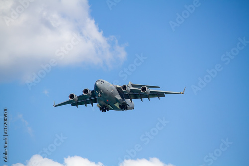 C-17 Globemaster III military transport aircraft in flight, viewed from below against a blue sky with a white cloud below the aircraft, showing its four jet engines and distinctive high-wing design.