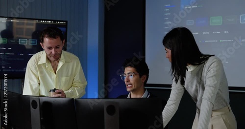 Adult female technology professional leans beside workstation, discusses software dashboard with two male colleagues, and gestures while team reviews digital system data during office collaboration.