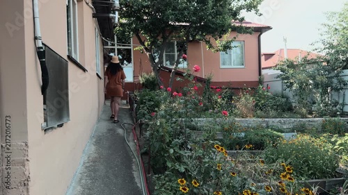 Unrecognizable man with long hair in bucket hat entering frame and walking away along path beside rural house wall and lush garden with blooming red roses leaving scene empty authentic rear view