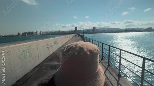 Unrecognizable traveler wearing beige bucket hat walking along concrete pier towards distant city skyline. Sunny day outdoor rear view tracking shot over sea water capturing freedom and solitude