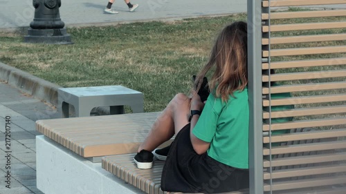 Unrecognizable young woman in green shirt relaxing on modern wooden bench in city park using smartphone with hair blowing in the wind side rear view casual outdoor lifestyle chilling alone texting