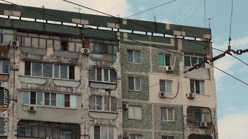 Weathered multi story residential apartment panel building facade with enclosed balconies and overhead trolleybus wires in post soviet Eastern European city under cloudy sky static establishing shot
