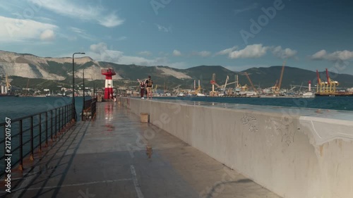Wide establishing shot of unrecognizable couple enjoying freedom on concrete pier in Novorossiysk seaport with red lighthouse, industrial cranes, sea water and mountains under cloudy blue sky