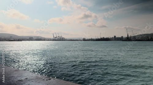 Wide establishing shot of rippled blue sea water surface with bright sun glare reflecting on waves in large industrial seaport bay with distant city skyline and cargo cranes under cloudy sky