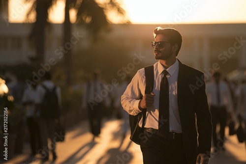 Young businessman walking outdoors wearing a suit with sunglasses during golden hour