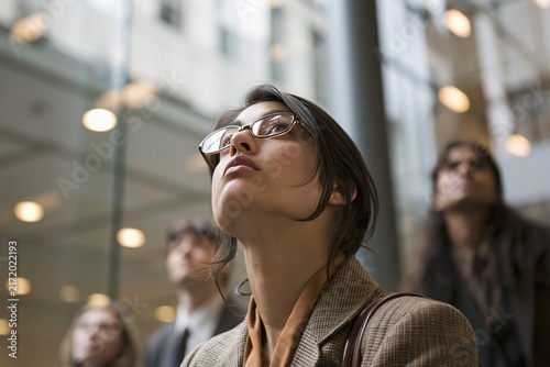 Young businesswoman wearing glasses looking upwards thoughtfully amidst a crowd in a modern building