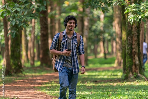 Young indian man walking through lush green forest trail wearing headphones and casual plaid shirt