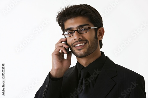 Young man with glasses and a black suit talking on a mobile phone against a plain white background