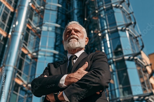 Confident senior businessman with gray beard standing outdoors with arms crossed before skyscraper