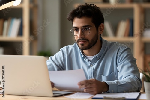 Focused man wearing glasses working on a laptop while reviewing paper documents at his desk