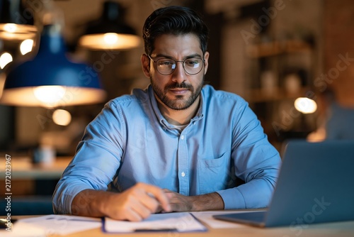 Focused man working late at night in an office with a laptop and documents on his wooden desk