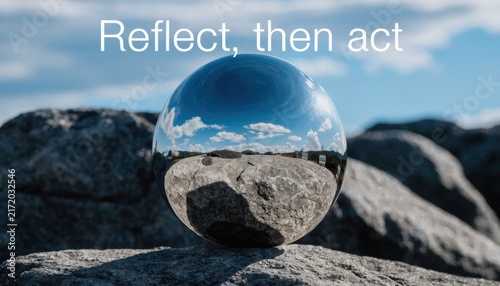 Polished chrome metal sphere reflecting clear blue sky and clouds, resting on a textured grey rock with white text overlay reading Reflect then act in a mountainous landscape.