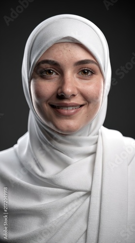 Front-facing headshot of a happy young Muslim woman with brown eyes and freckles wearing a white textured hijab, looking at the camera on a neutral studio background.