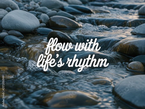 Close-up shot of a gentle mountain stream flowing over smooth grey river stones and pebbles with white script typography reading flow with life's rhythm in a serene natural setting.