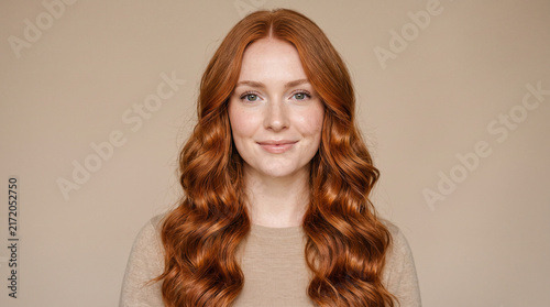 Smiling Young Woman with Long Copper Red Wavy Hair Posing Against Beige Background