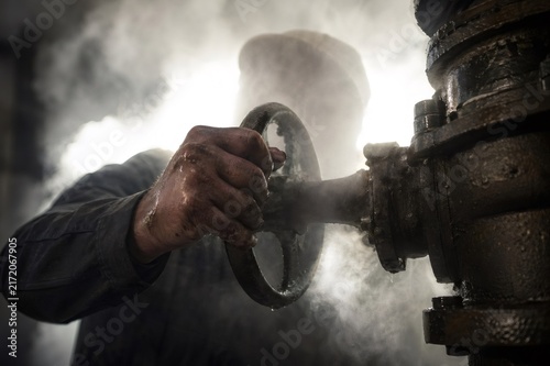 Worker hand covered in grime and water turning a large metal valve on a pipe, releasing steam and showing the harsh realities of manual labor and industrial processes