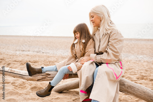 Mother holding child daughter sitting at log at beach wearing similar beige coats spending time together. Motherhood. Autumn season.