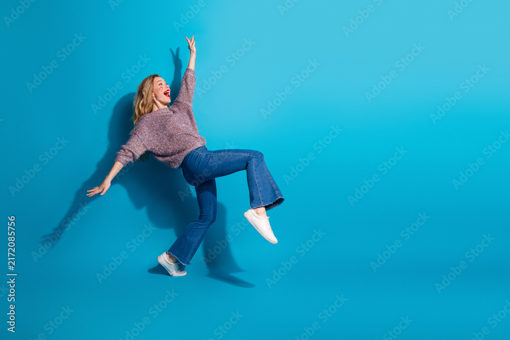 Fototapeta premium Young woman in casual sweater jeans jumps with joy against blue background