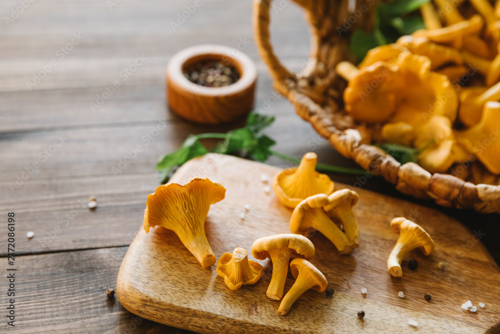 Obraz premium Fresh chanterelle mushrooms on a wooden board and in a basket on a dark table, rustic harvest scene, healthy cooking ingredients, organic forest food, autumn season. Top view, flat lay, copy space.