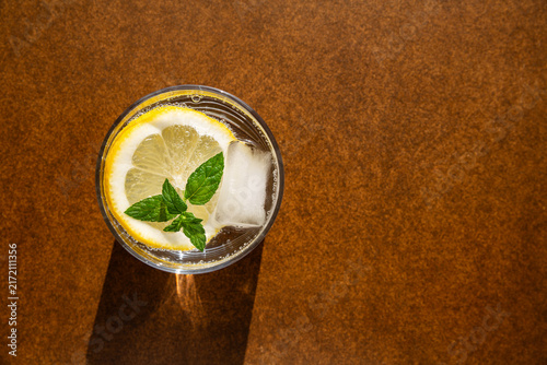 Un verre transparent rempli avec de l'eau pétillante et une rondelle de citron jaune, des feuilles de menthe et un glaçon, fond brun, vue du dessus, boisson fraîche, espace libre à droite