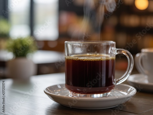 Close up of a steaming glass mug of black coffee on a white saucer placed on a wooden table in a blurry cafe interior with soft bokeh background and rising steam.