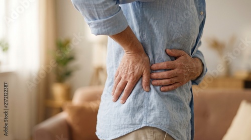 Horizontal photo of an adult person standing and holding their lower back in pain, spine issues concept