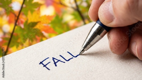 Person writing the word Fall on paper with autumn leaves background