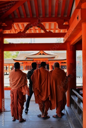 Fotografía Back view of Buddhist monks in traditional orange saffron robes visiting the iconic red wooden corridors of Itsukushima Shrine in Miyajima, Japan