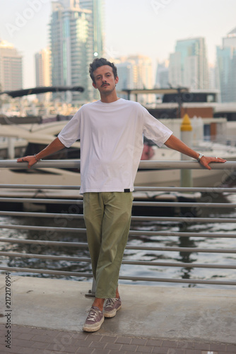 Blank white oversized t-shirt mockup on a young male model, modern city marina background with yachts and skyscrapers