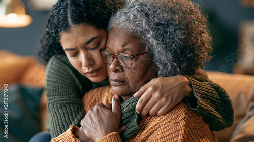 A daughter embraces her elderly mother who is feeling unwell. Holding her mother's hand giving support and consoling her while sitting on sofa in the living room at home