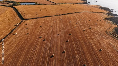 Drone camera orbit over coastal farm fields with hay bales bathed in evening sun, revealing rugged cliff edge and calm sea below