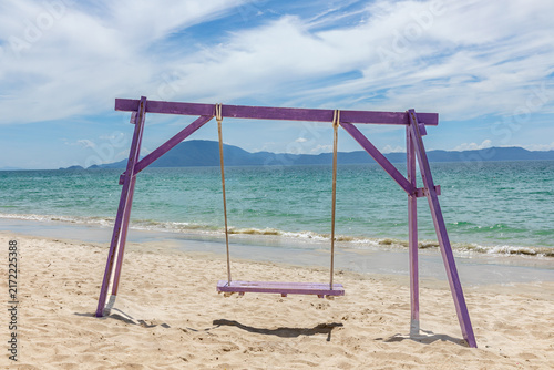 Canvastavla A purple wooden swing set stands on a sandy beach, with clear blue water gently lapping at the shore and distant mountains visible under a bright, partly cloudy sky