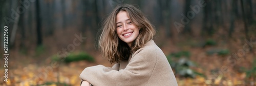 Smiling young woman sitting in an autumn forest with a coat and fallen leaves for seasonal portrait and nature background