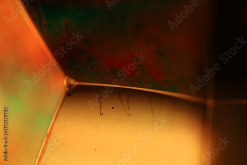 Geometric liquid film structure with warm orange glow and green iridescent shadows. Crisp specular lines outline a geometric liquid film corner in low-key macro perspective.