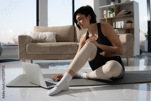 Woman using laptop for online fitness class while sitting on yoga mat at home