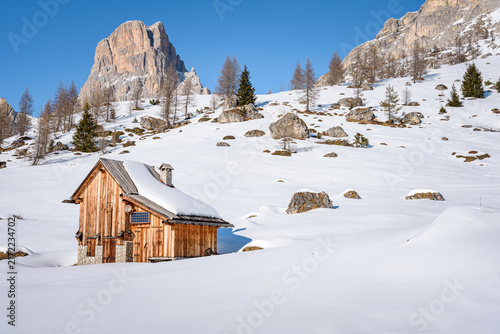 Фотография Lonely wooden mountain hut on a snowy slope at the foot of rocky peaks in the European Alps on a sunny winter day